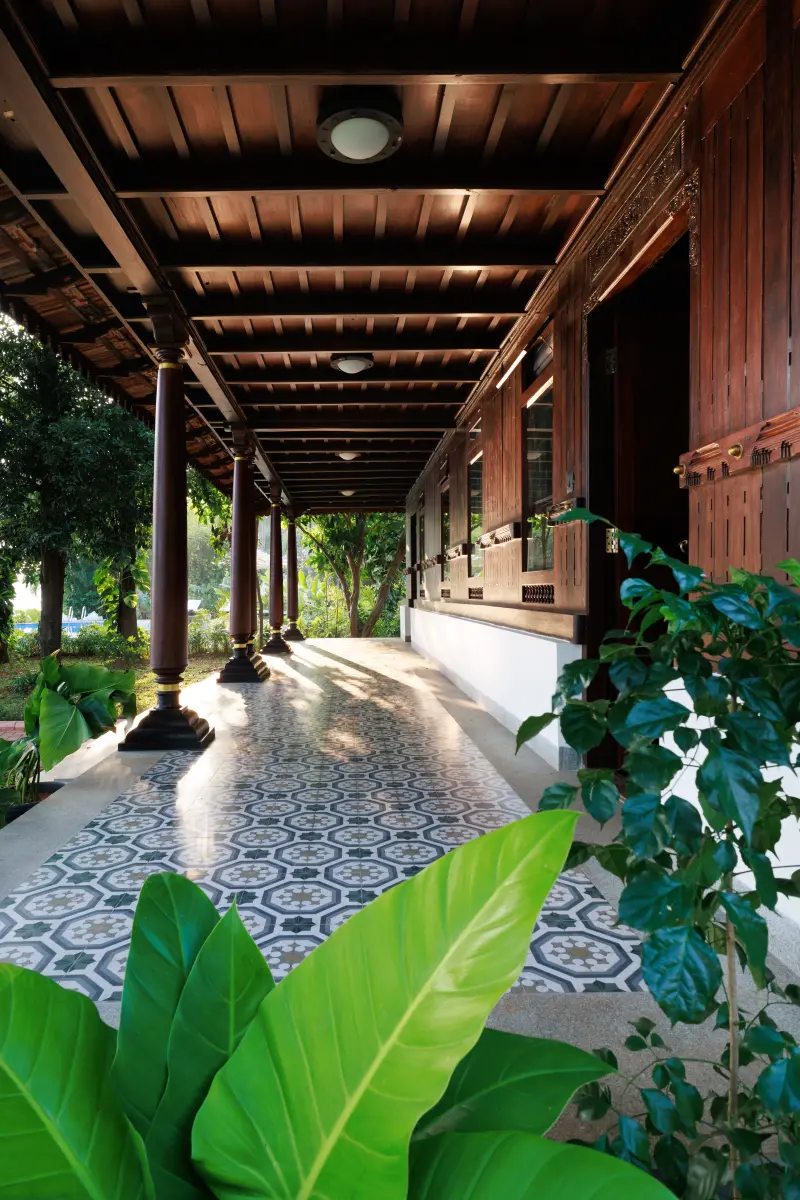 Heritage Room veranda corridor at Vedic Village Kodungallur with carved teak columns, traditional patterned tile flooring and tropical garden foliage