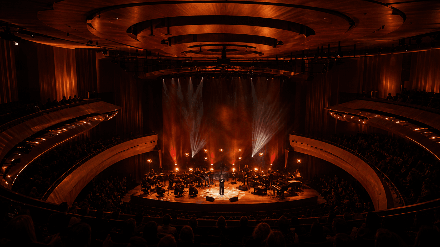 An orchestra and vocalist perform under warm lighting in a full concert hall.