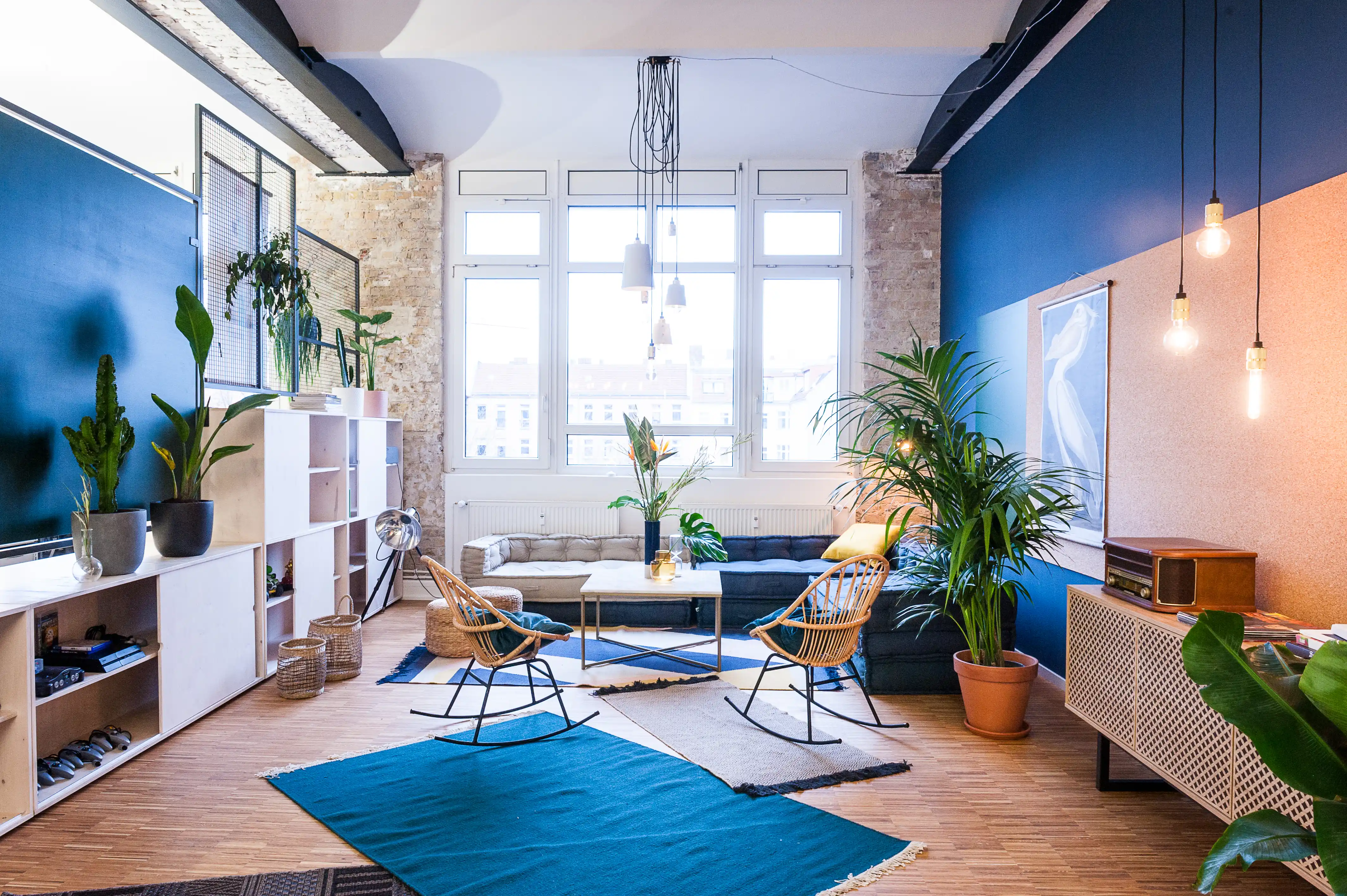Loft lounge at INTERMATE x TRUEMATES with a deep blue wall, exposed brick, large windows, a navy sofa, rattan rocking chairs, layered blue rugs, white shelving and a sideboard with mesh doors.