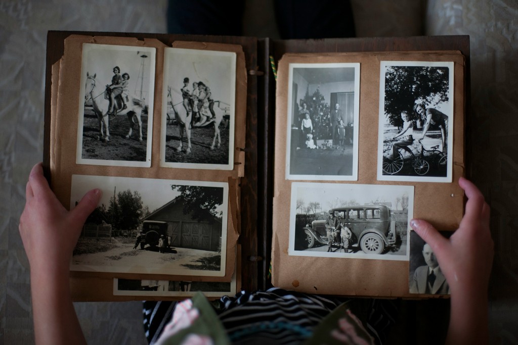 A person is holding an old family photo album featuring vintage black-and-white photographs. The images include children riding horses, a family gathering around a Christmas tree, kids on a tricycle, and a classic car with people standing beside it. The nostalgic collection captures moments of joy and history, evoking a sense of heritage and family memories.