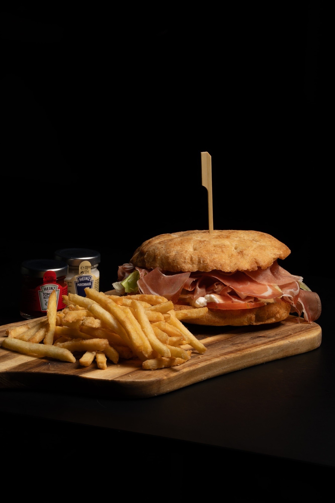 Panino con crudo, stracciatella e pomodoro accompagnato da patatine e barattolini di salse, servito su tagliere in legno