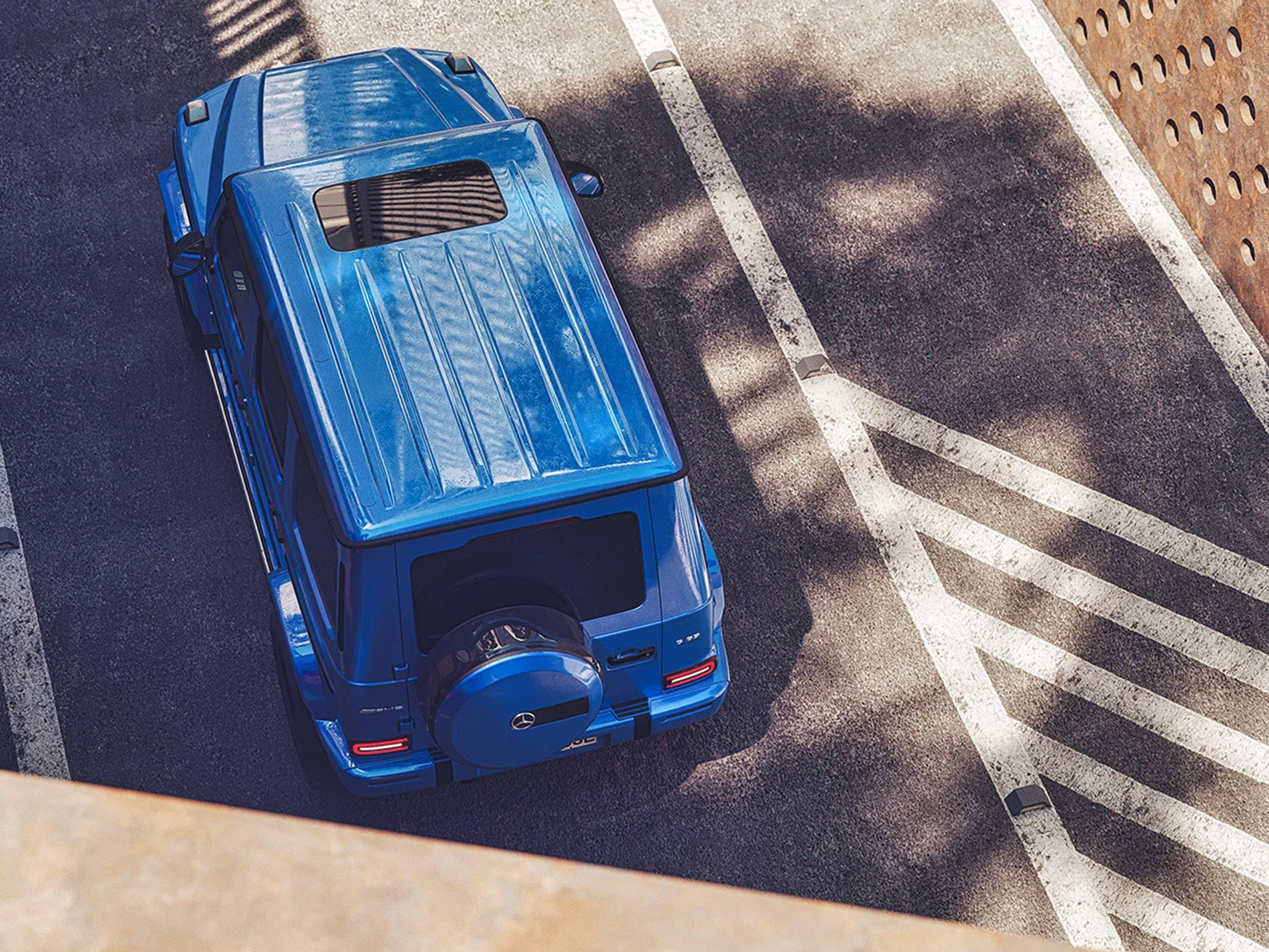 Vista aerea di un SUV Mercedes Classe G blu parcheggiato tra strisce bianche stradali, con dettagli del tetto e design squadrato iconico.