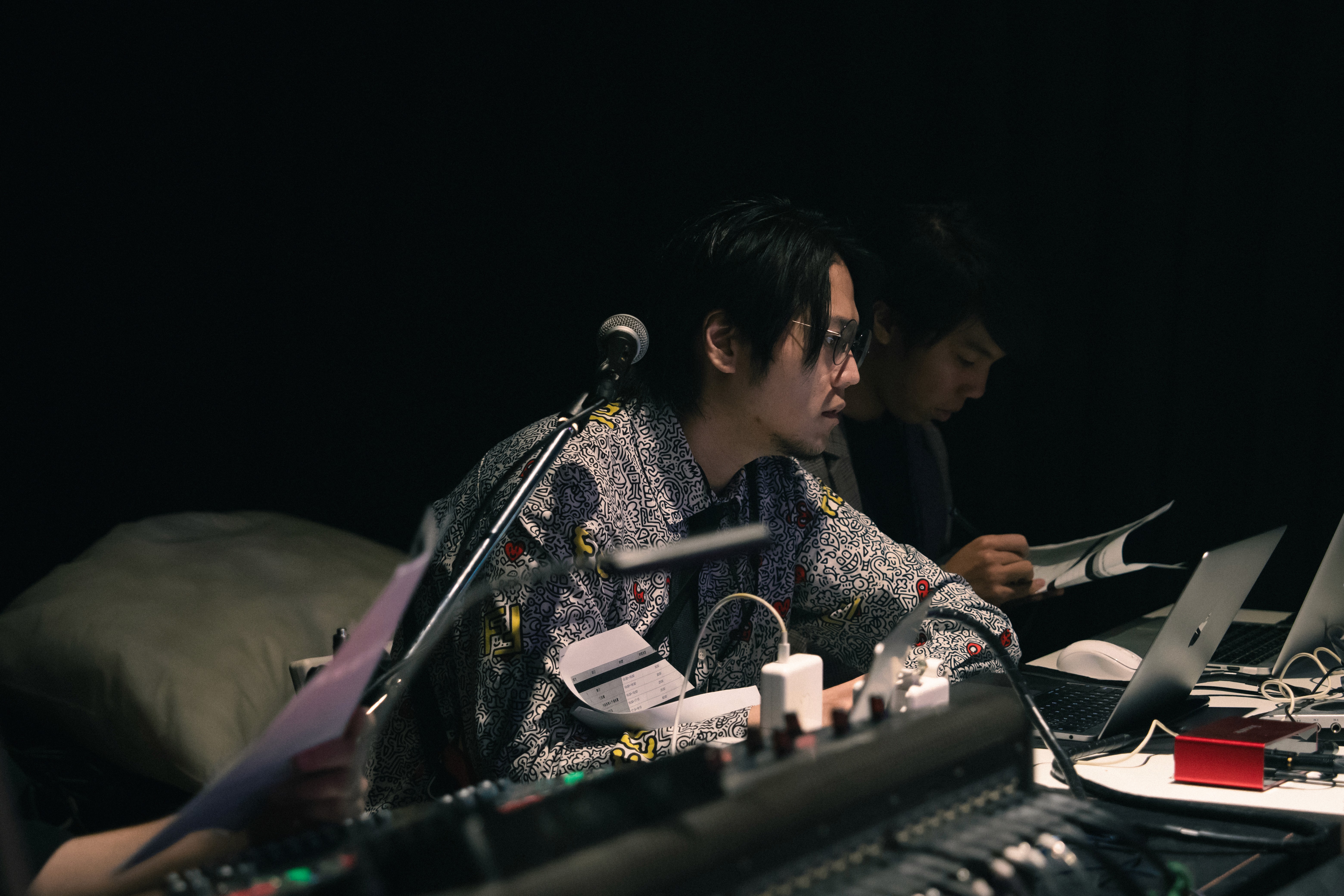 Two camera operators in a dimly lit studio adjust a large video camera. They wear headsets and caps, focusing intently. Mood is professional and focused.