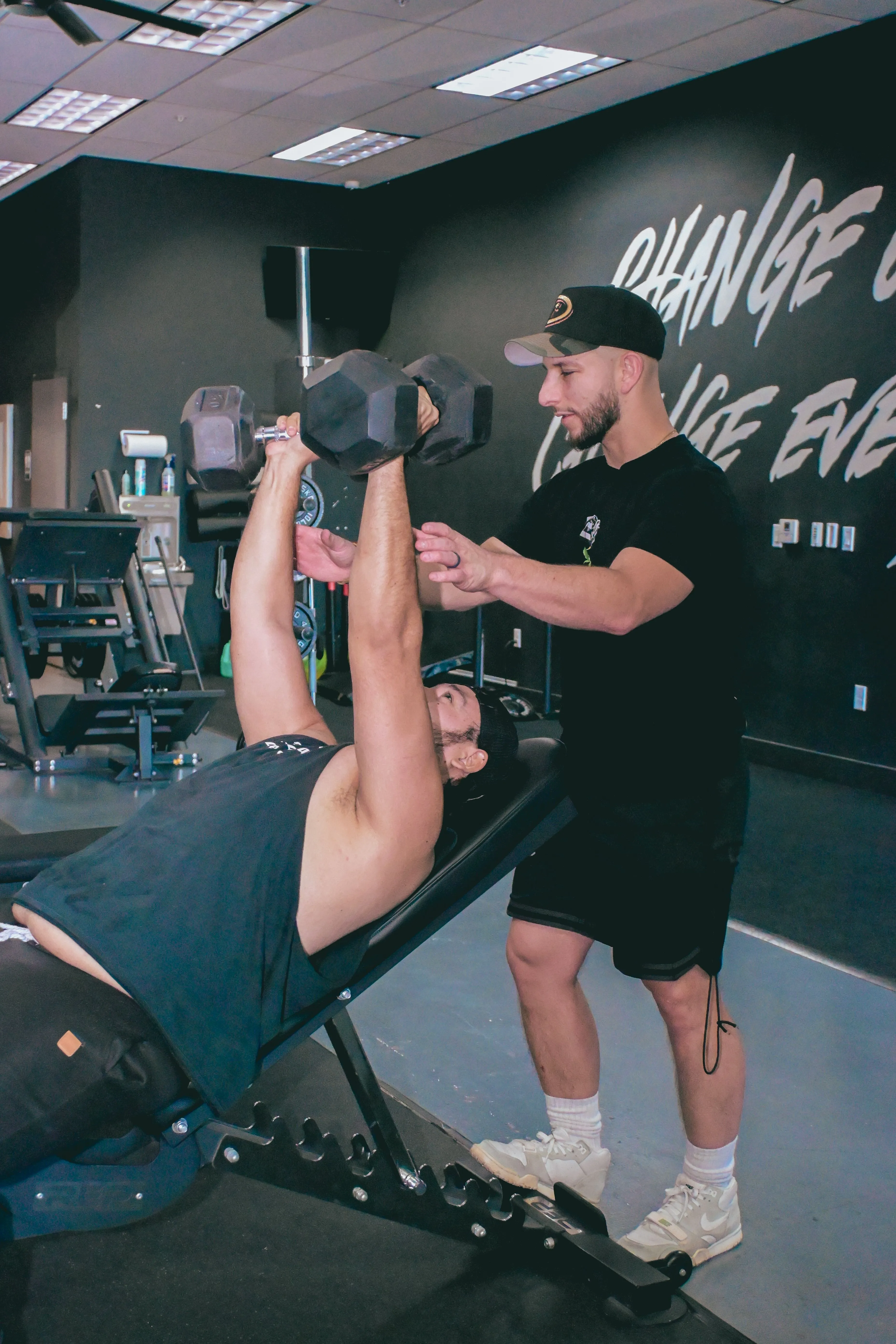 A person lies on a bench performing a dumbbell press, while another person stands nearby in gym attire, offering guidance in a modern fitness facility with motivational wall text.