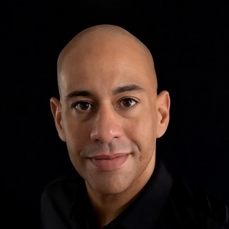 Smiling bald man in yellow shirt against black background.
