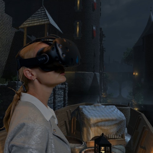 Egy személy, aki egy virtuális valóság headsetet visel egy középkori témájú környezetben, ahol kőépületek és egy híd látható a háttérben.