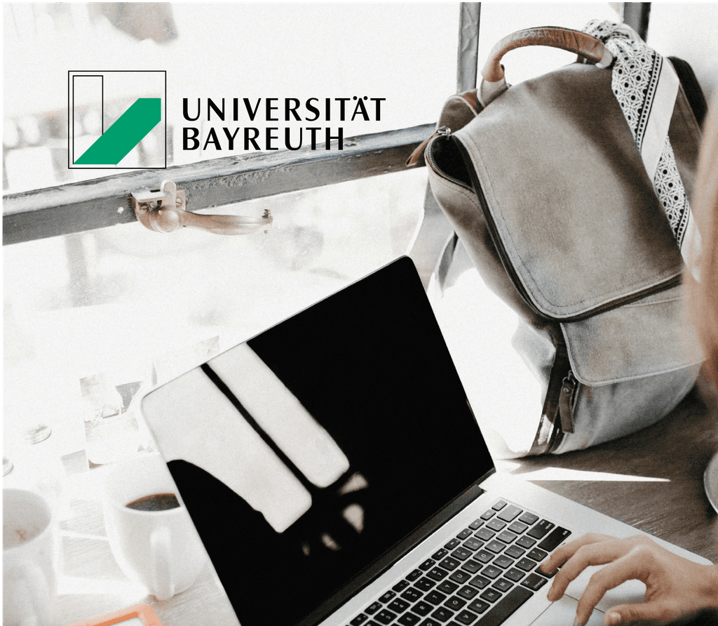 Eine Frau sitzt mit ihrem Laptop an einem Schreibtisch in einem Unicafé und erledigt Aufgaben, auf dem Bild befindet sich das Logo der Universität Bayreuth