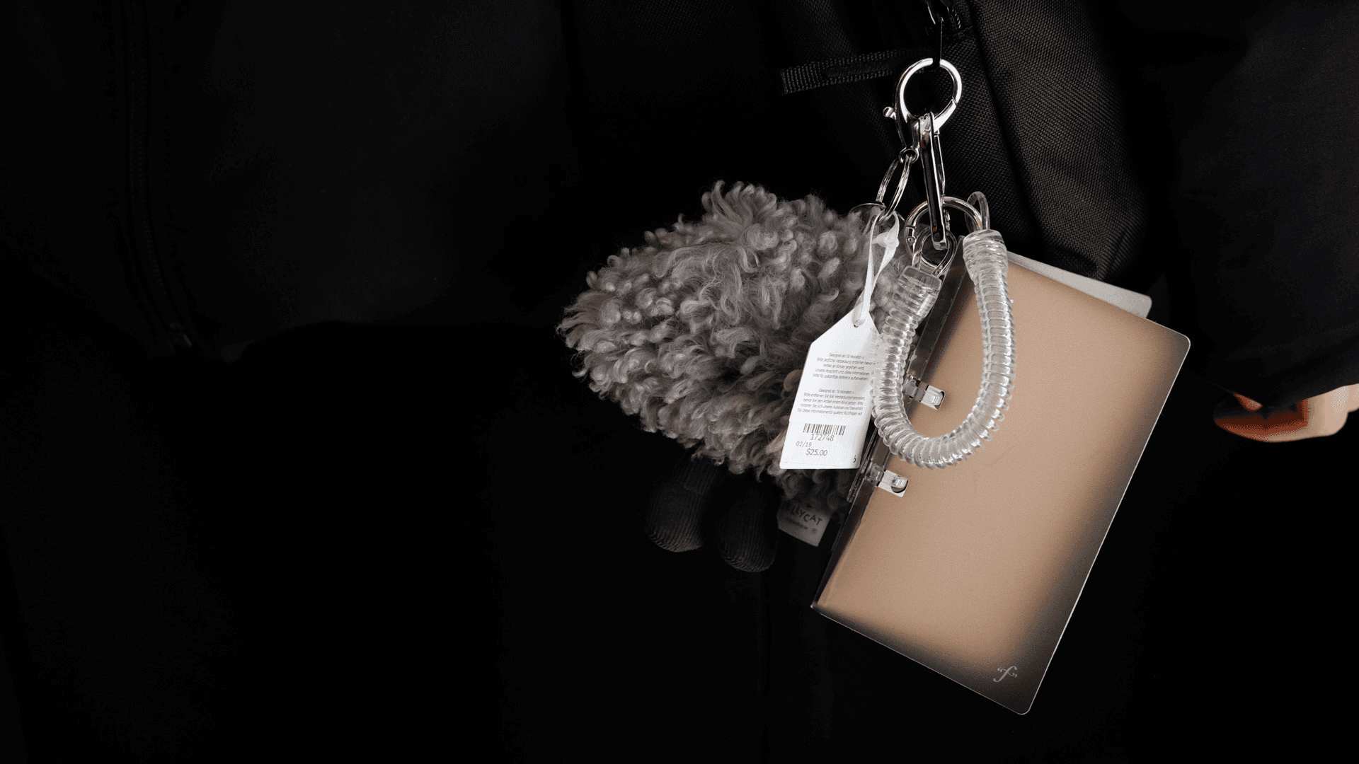 A light beige or tan clutch bag hangs from a silver metal keychain loop. A fuzzy gray item is attached to the bag, and a white tag with black text is attached to the loop. The background is dark, and the bag is partially visible against a black surface.