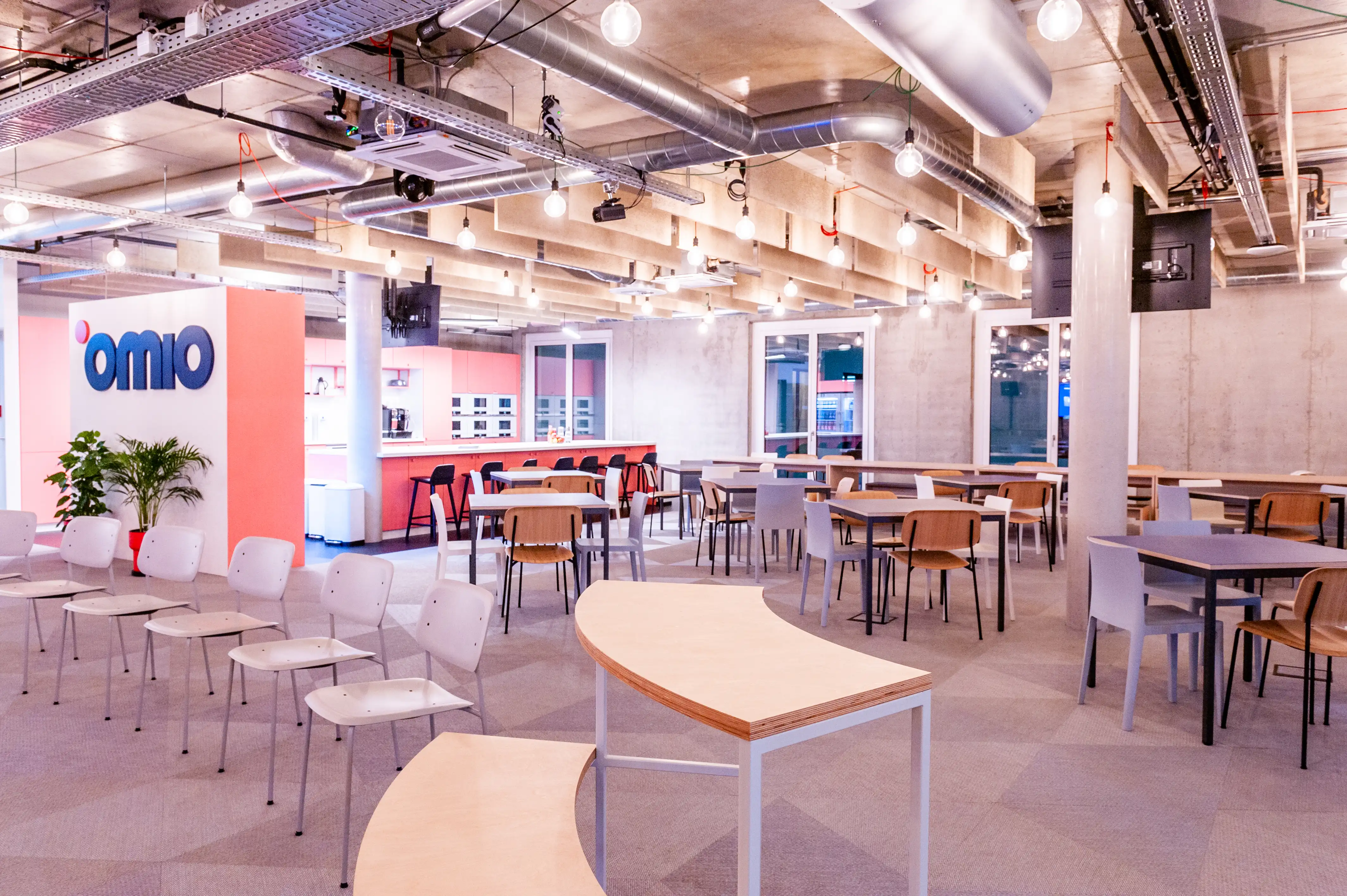 Large canteen at OMIO with mixed seating of white chairs and plywood tables, a curved plywood bench, the coral kitchen counter with bar stools and the blue Omio logo on a white wall.