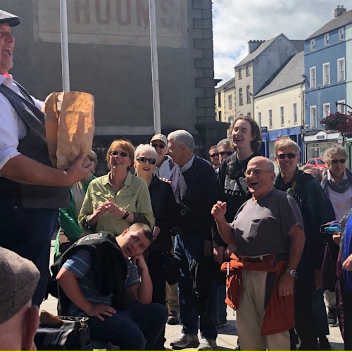 在阳光明媚的一天，一名男子手持纸袋，在市中心广场上对着一群微笑的围观者讲话。