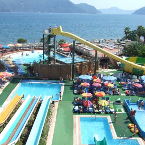 A water park with various slides, pools, and people under colorful umbrellas situated near a seaside with mountains in the background.