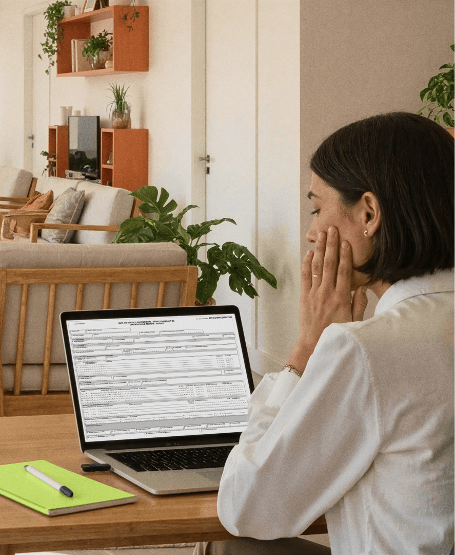 Profissional em área de recepção de clínicas analisa no notebook uma guia TISS, na Tivita; na mesa há um caderno verde e caneta, e ao fundo aparecem poltronas de madeira e plantas