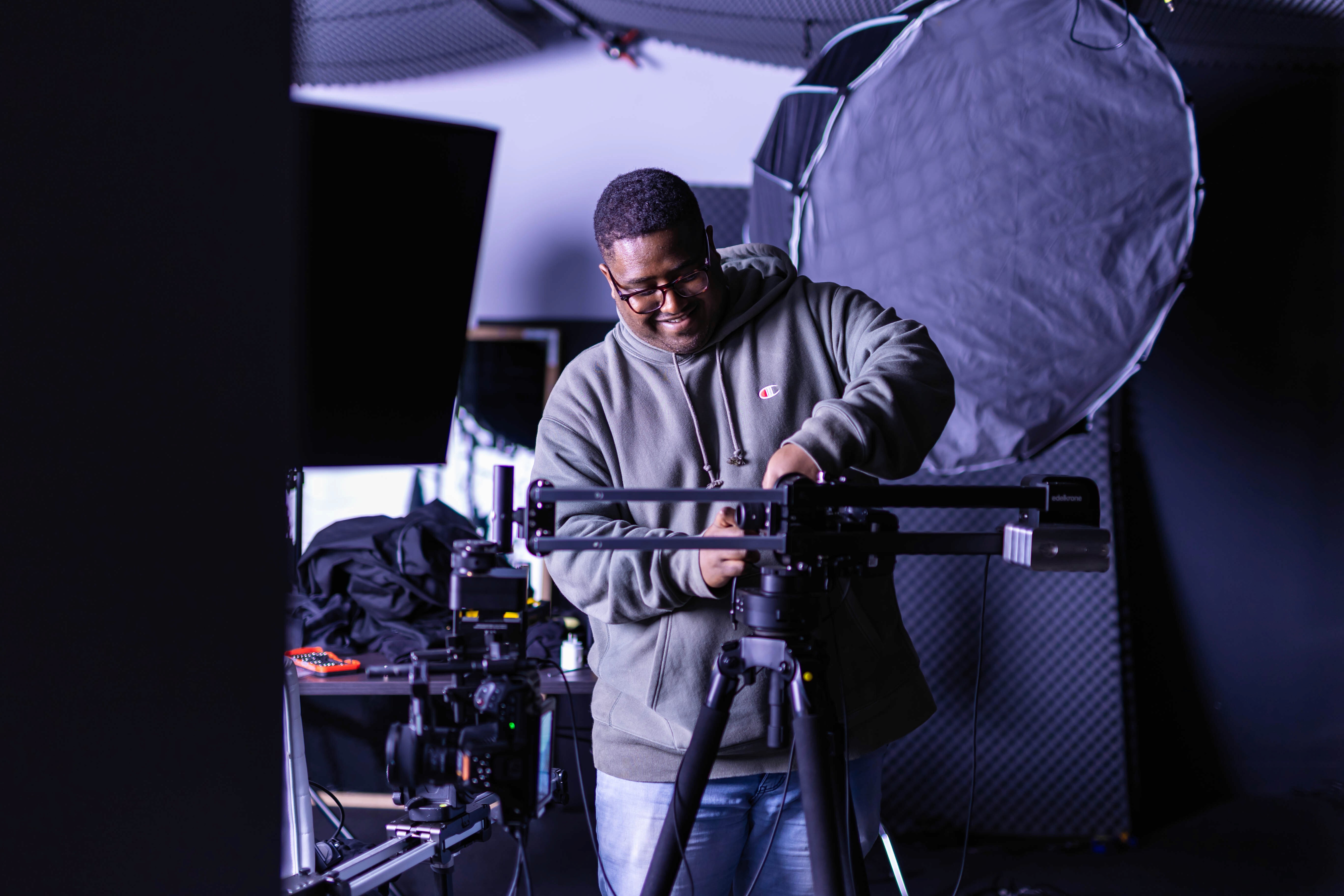 Two filmmakers operate a large camera on a dimly lit set. One wears headphones, both focused, suggesting a serious, creative atmosphere.