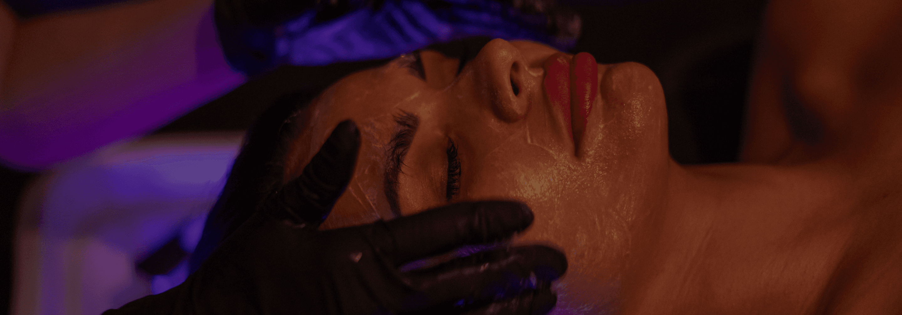 A person receives a facial treatment with gloved hands applying a skincare product, illuminated by soft purple lighting, emphasizing their serene expression and the luxurious spa environment.