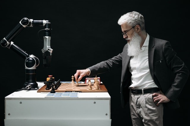 A bearded man plying chess with a robot arm.