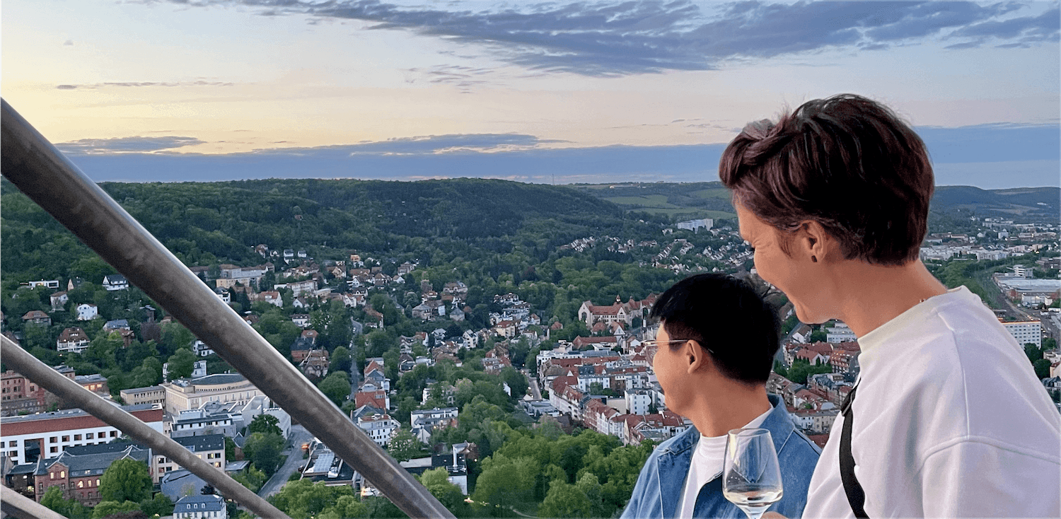 Zwei Personen stehen auf einer Aussichtsplattform mit Metallgeländer und blicken auf eine Stadt mit vielen Gebäuden und viel Grün in der Dämmerung