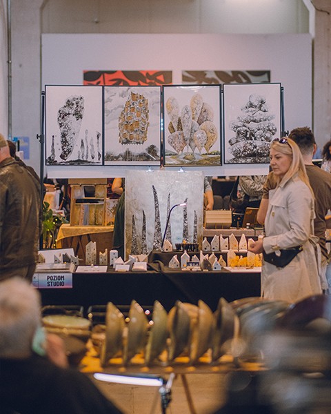 Zdjęcie przedstawiające ludzi przechądzących między stoiskami. W tle obrazy i ceramika.