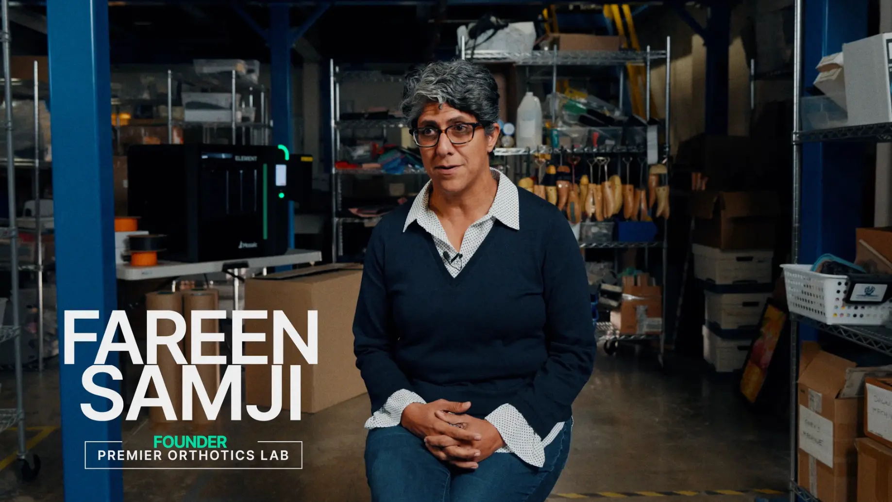 Fairen Samji, founder of Premier Orthotics Lab, sits in a workshop lab with tools and shelves behind them, with "FAREEN SAMJI," "FOUNDER," and "PREMIER ORTHOTICS LAB" shown on screen, supporting a medical device marketing strategy.
