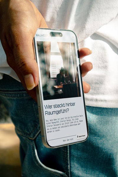 Mockup eines iPhones in einer Hand, auf dem die Raumgefühl-Website geöffnet ist. Sichtbar ist die Seite mit einem Bild einer Person am Schreibtisch und der Überschrift ‚Wer steckt hinter Raumgefühl?
