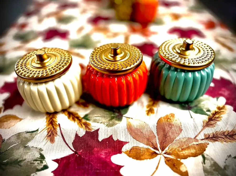 Three decorative, ribbed ceramic jars in cream, orange, and teal with gold lids are elegantly displayed on a floral tablecloth, adding a touch of autumn warmth and charm, perfect for a stylish home decor in Bradenton, FL.