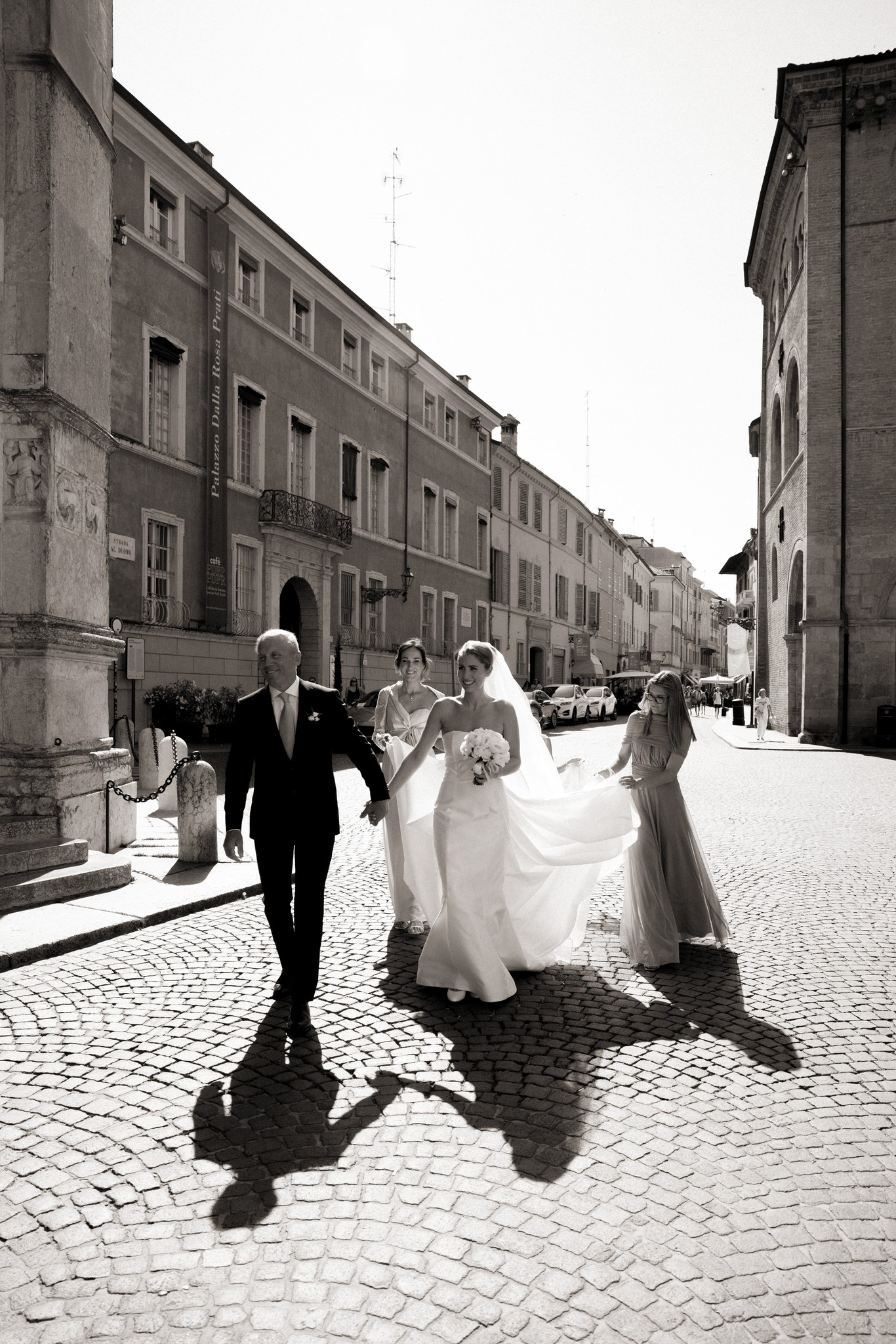Sposi camminano in piazza storica, strascico bianco segue in luce naturale, corteo matrimoniale a Villa Malenchini
