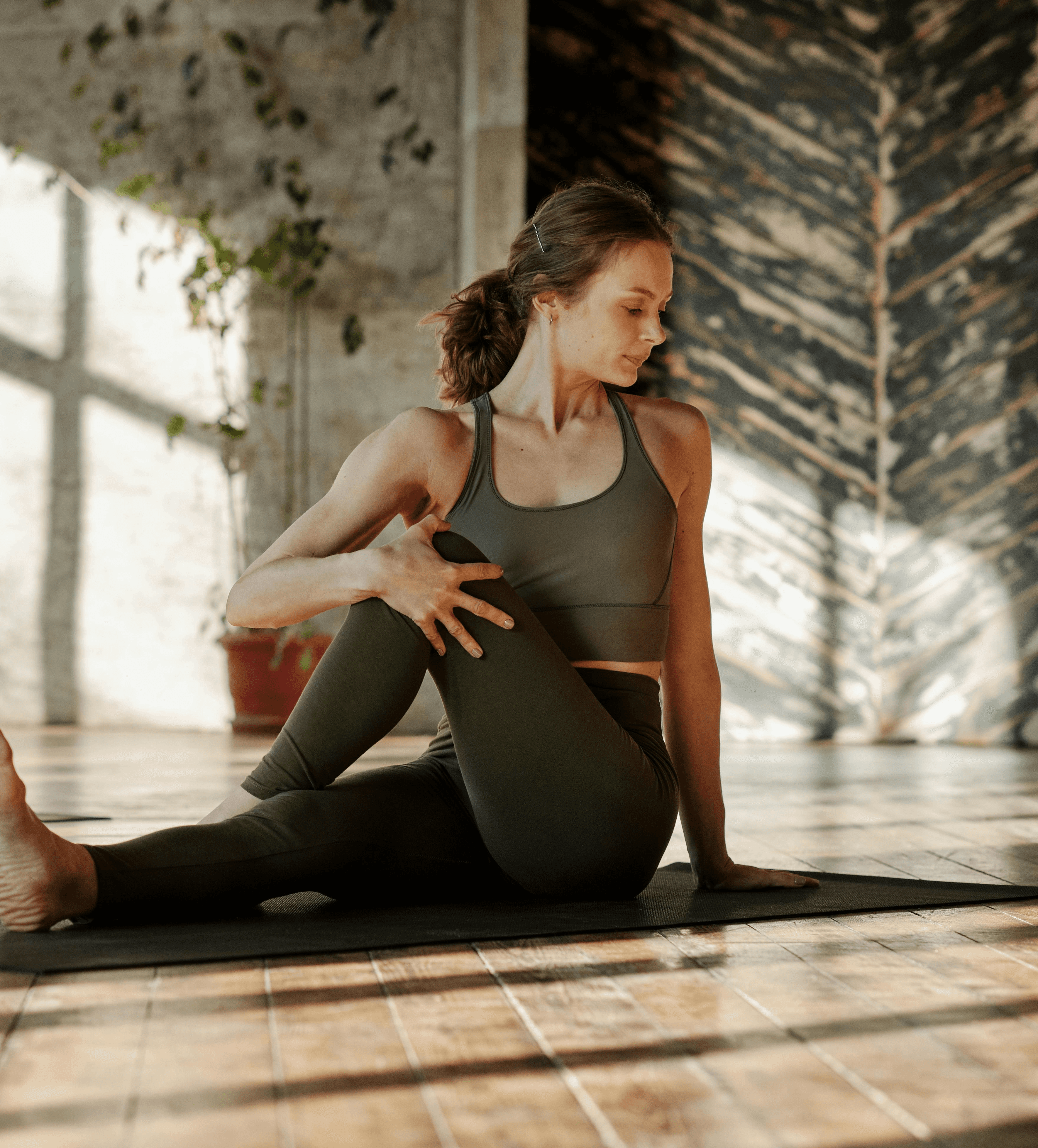 A woman in a yoga pose on a mat in a sunlit room with wooden floors. She wears athletic wear, looks focused, and the setting is peaceful with greenery in the background.