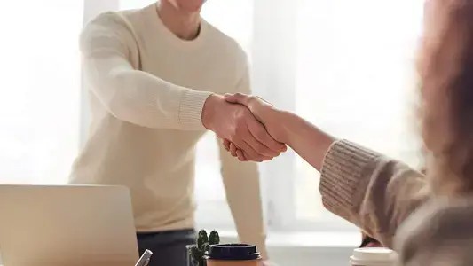 Two people are engaged in a handshake within a casual office setting, symbolising the striking of a business deal or agreement.