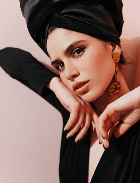 A woman in a black dress and turban, exuding elegance and sophistication, stands confidently against a neutral background.