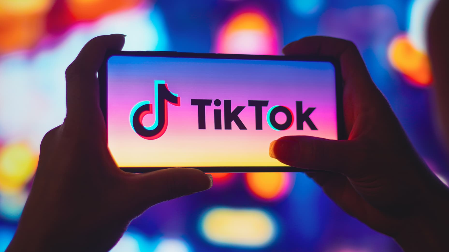 Hands holding a smartphone displaying the TikTok logo on a vibrant, multicolored background. The logo stands out with a sharp contrast against blurred neon lights.