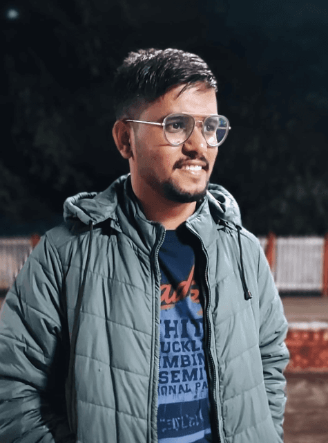 Portrait of a smiling man wearing glasses, a green jacket, and a graphic t-shirt, captured outdoors at night with a blurred background