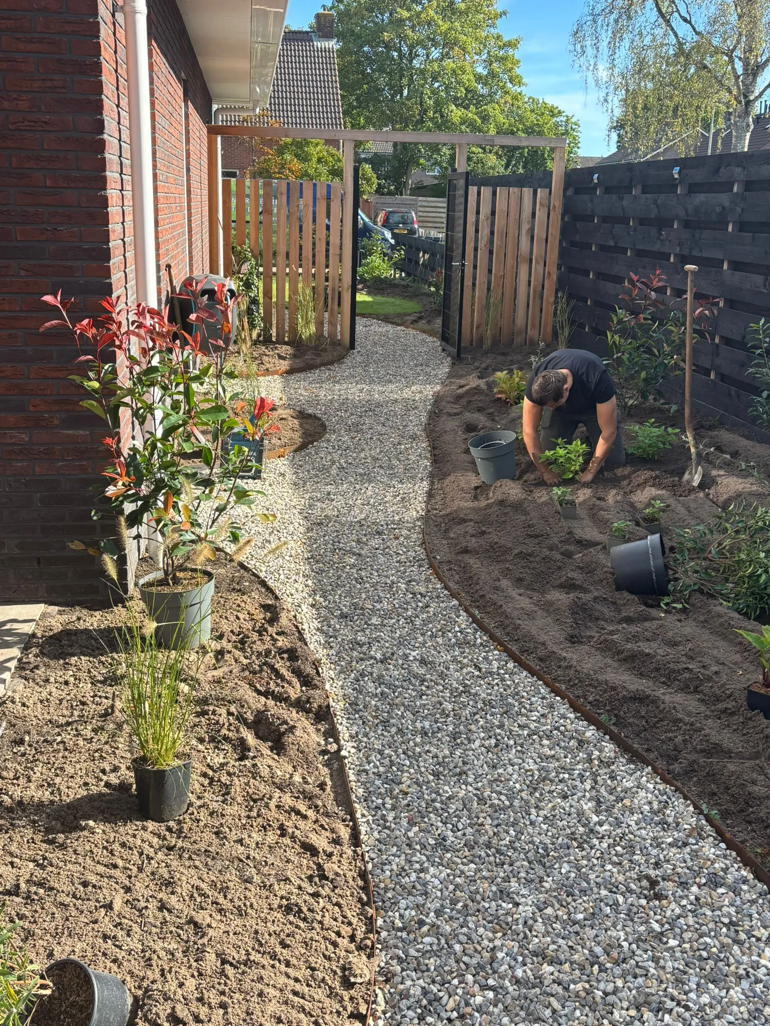 Tuinpad van kiezels met houten poort, links jonge planten in de grond, rechts hovenier die planten verzorgt bij zwarte schutting.