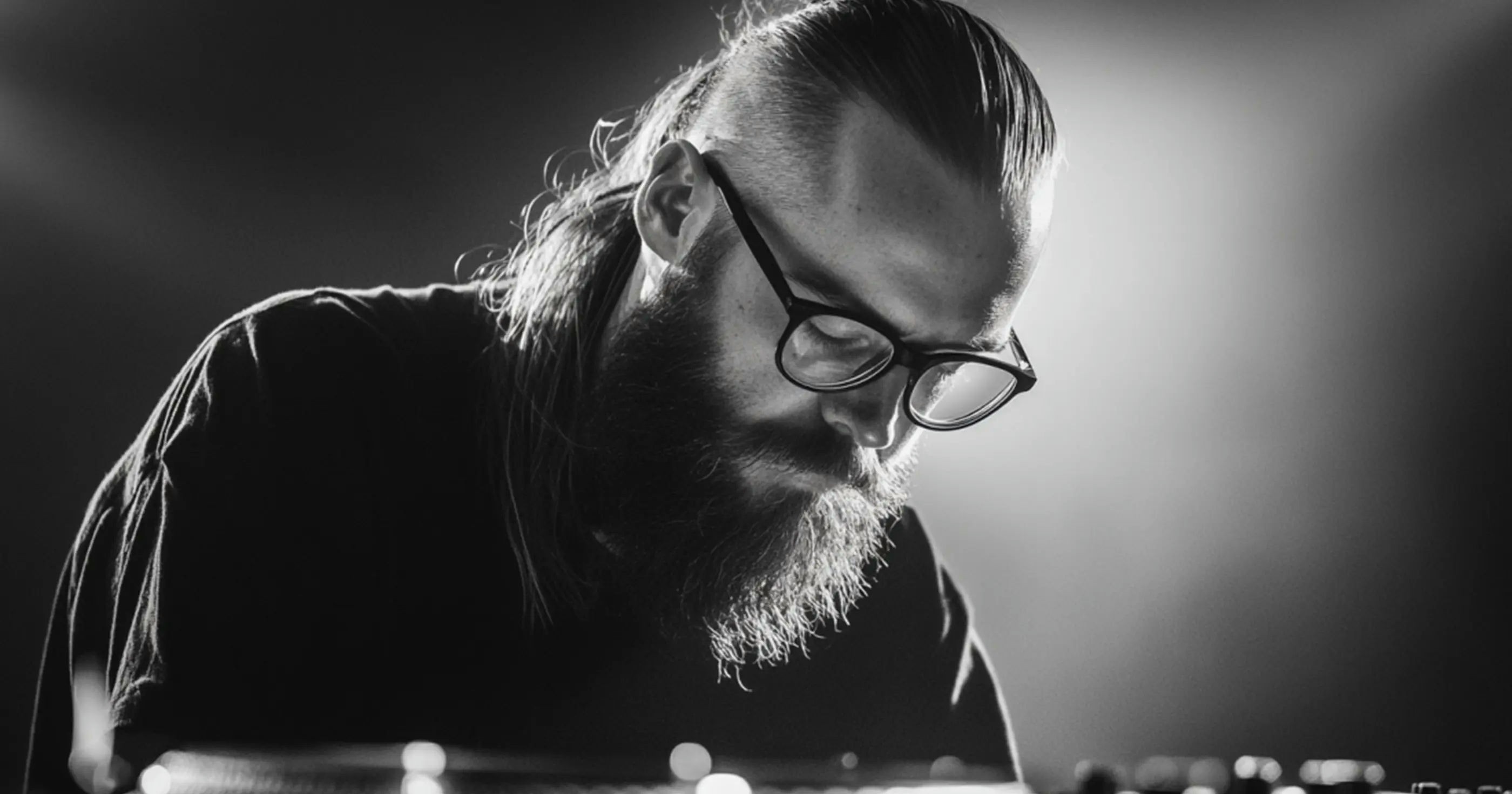 Black and white close-up of Skrillex with glasses and a beard, focused during a live performance.