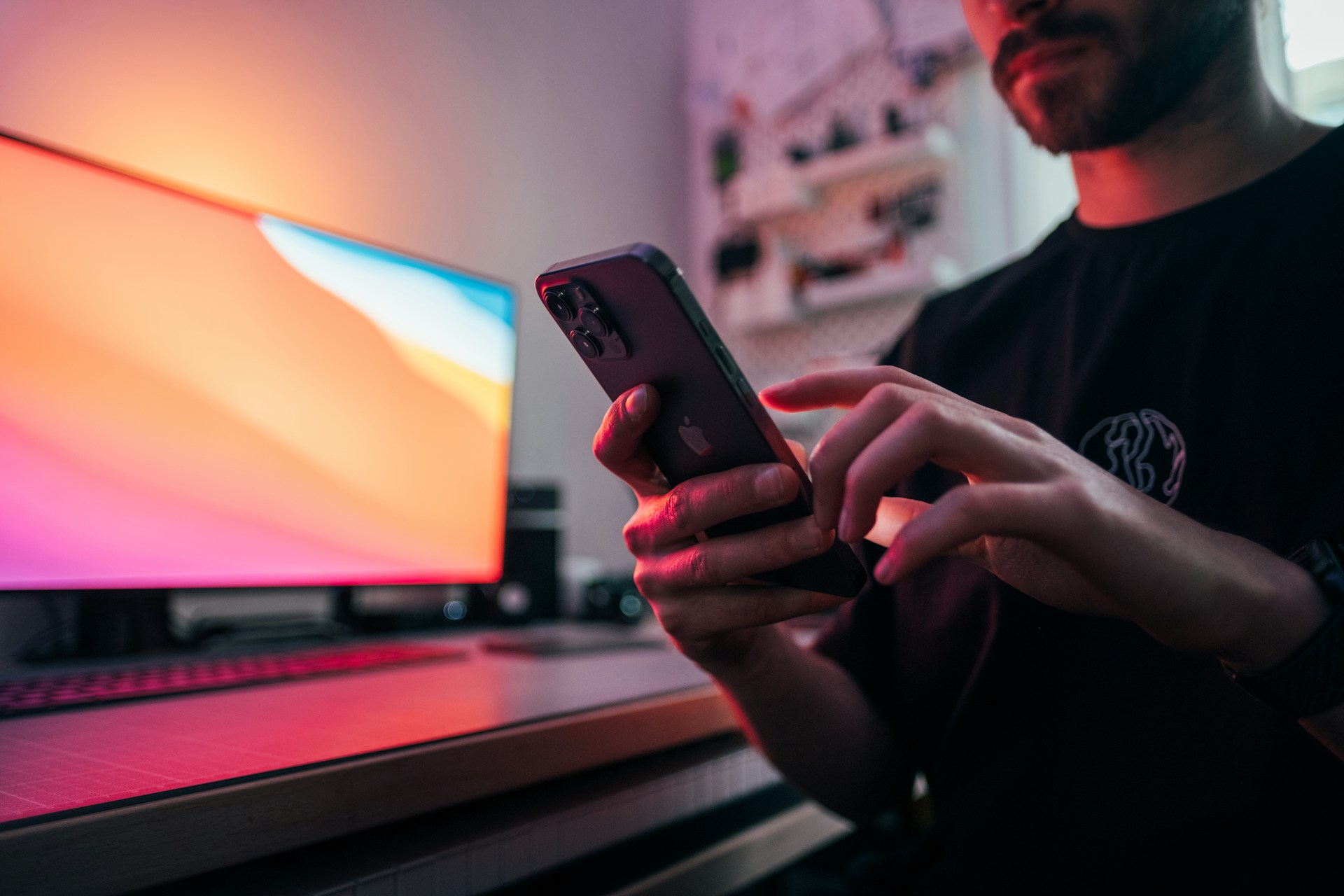 A person's hands are holding a dark smartphone with a black case, interacting with the screen in a dimly lit room with an orange-and-red glowing monitor in the background.