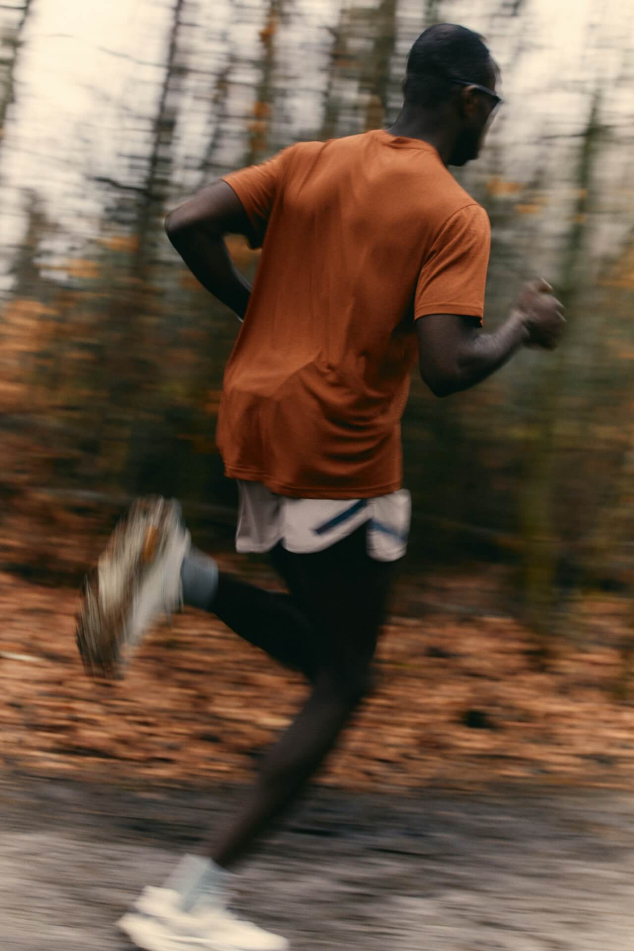A person in motion, wearing a brown shirt and white shorts, jogs through a blurred forest path. The scene conveys speed and energy.