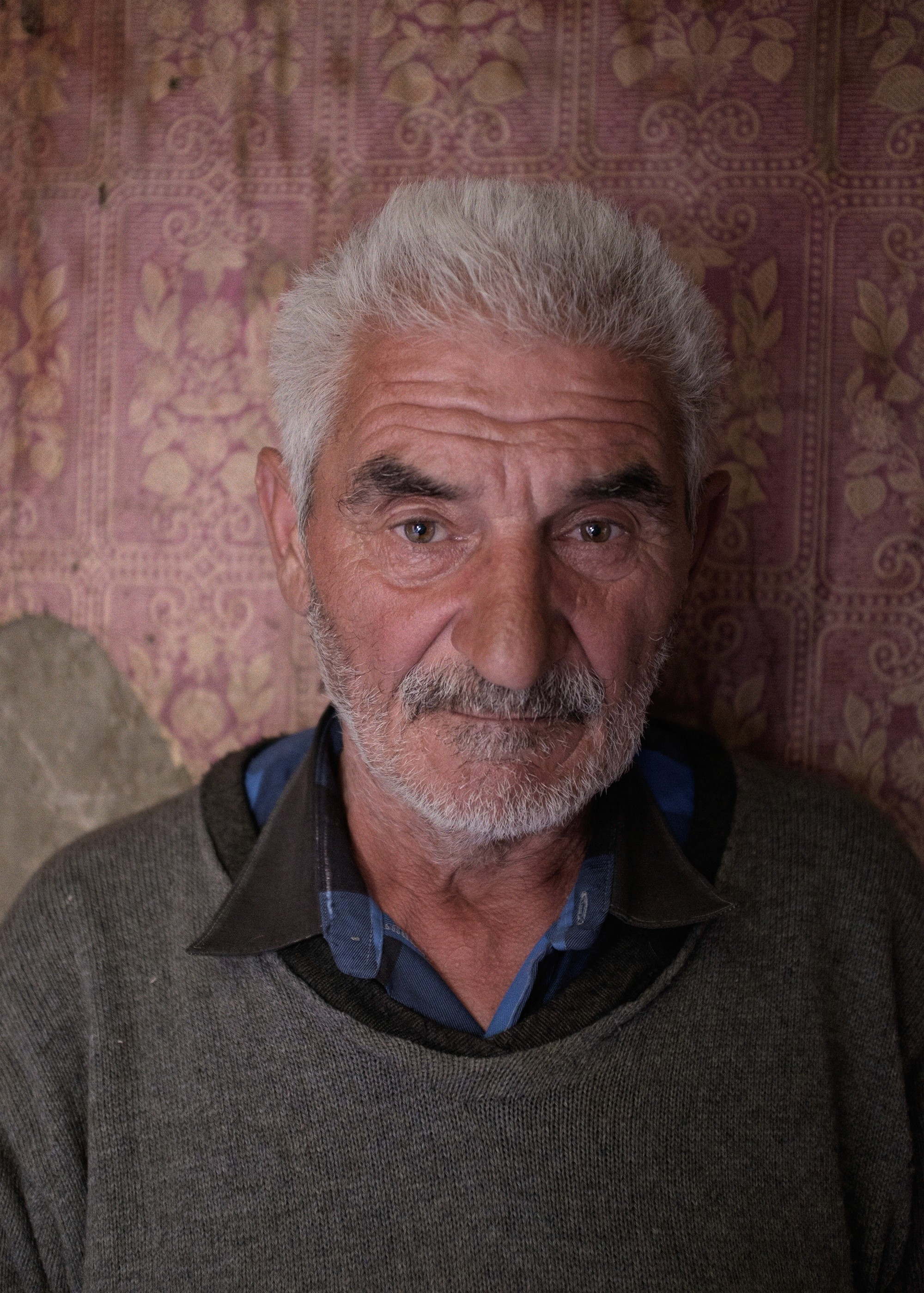 Portrait of Rezo, an elderly Georgian man with white hair and a trimmed beard, standing indoors against a worn floral-patterned wall