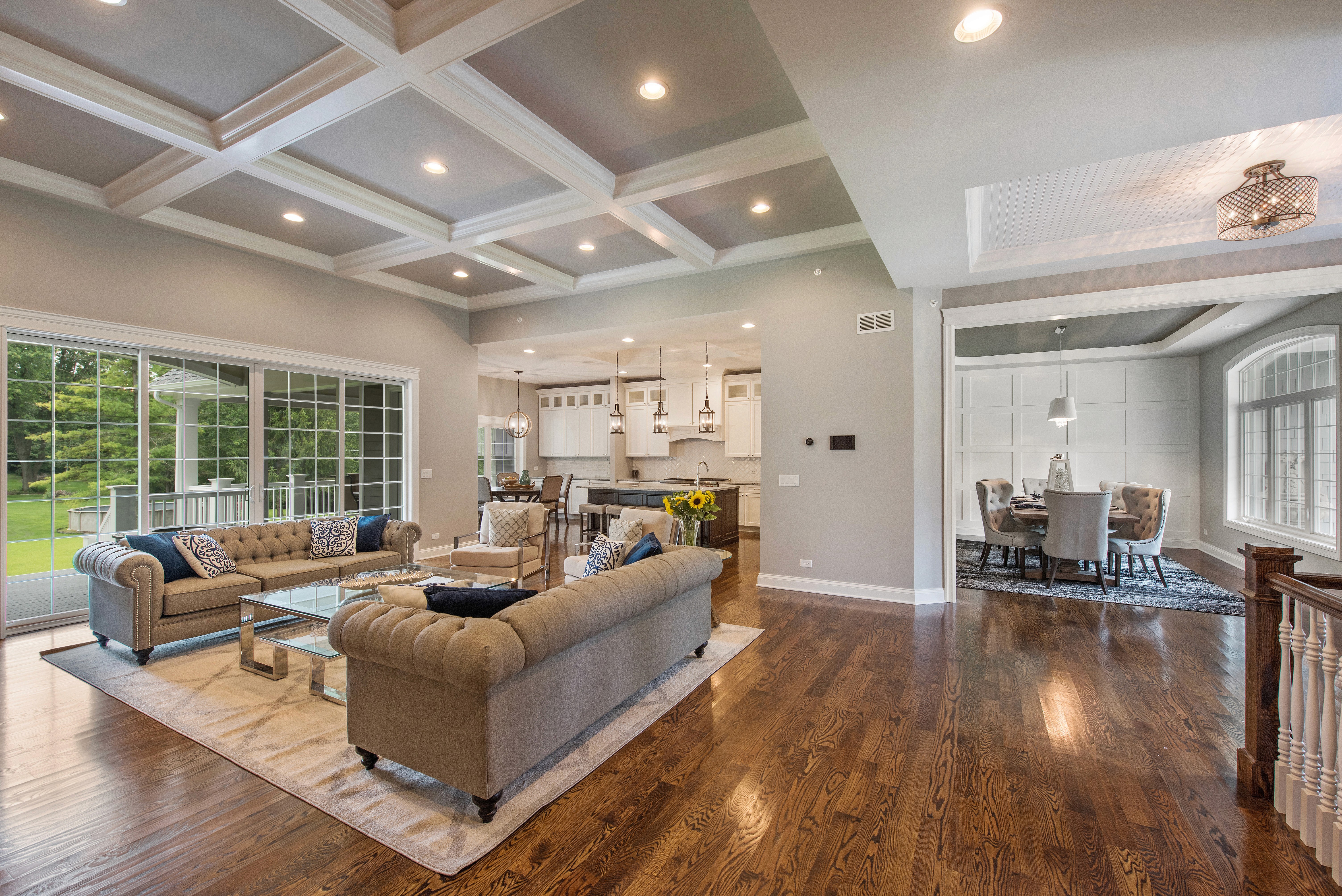 An open layout living room with furniture, coffered ceiling, a large sliding door, and stained hardwood flooring connecting to a kitchen and dining space