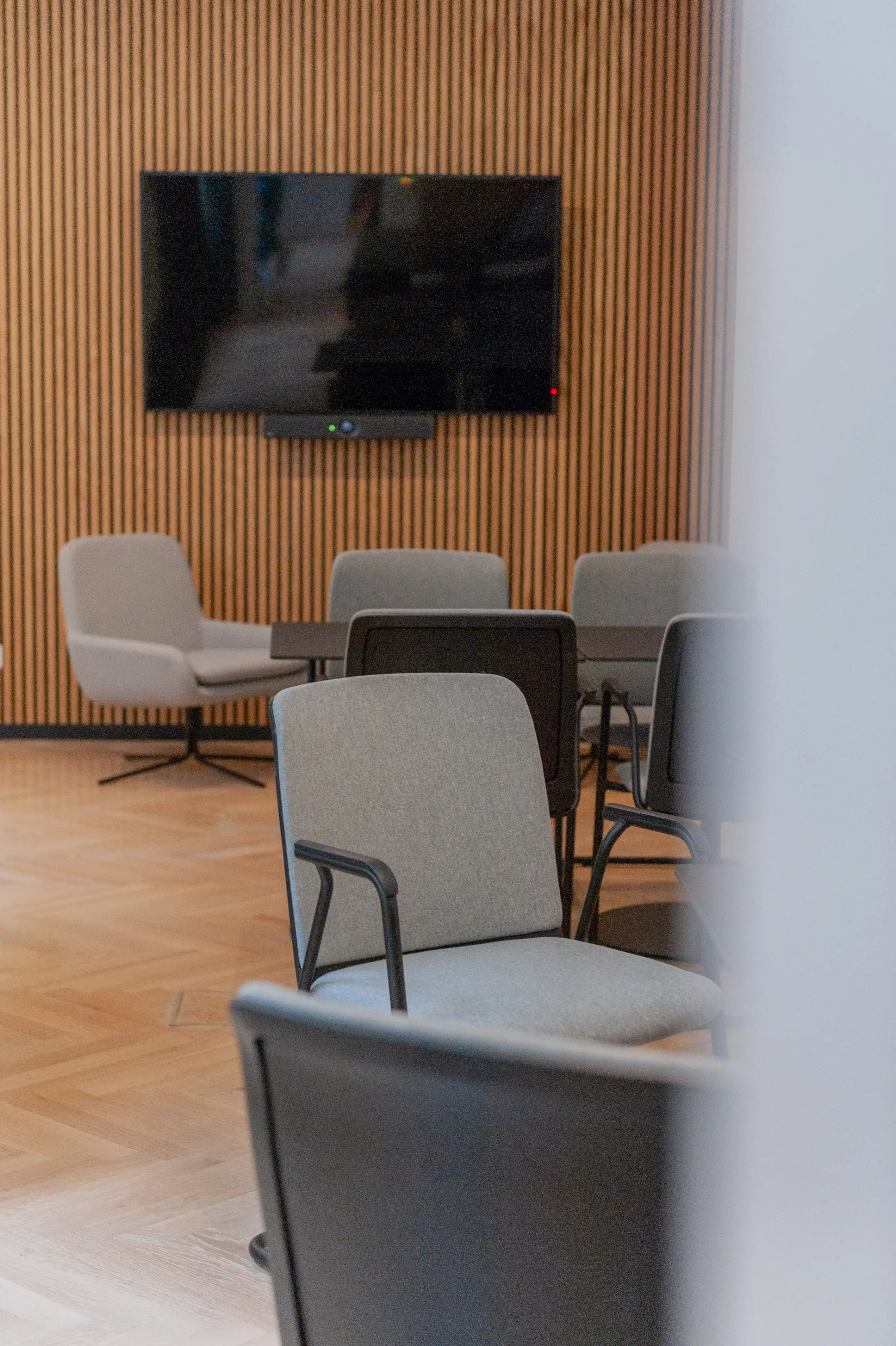 Meeting area at AI START-UP BERLIN with grey upholstered conference chairs, a wall-mounted TV with video bar on a wooden slat wall and a grey swivel armchair in the background.