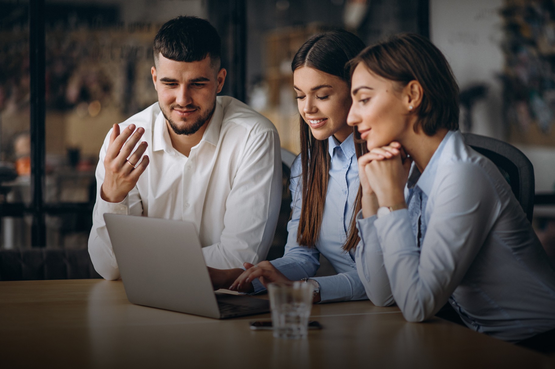 Three people in a modern office setting gather around a laptop, engaged in a collaborative discussion, with a glass of water on the wooden table, reflecting a professional and interactive work environment.