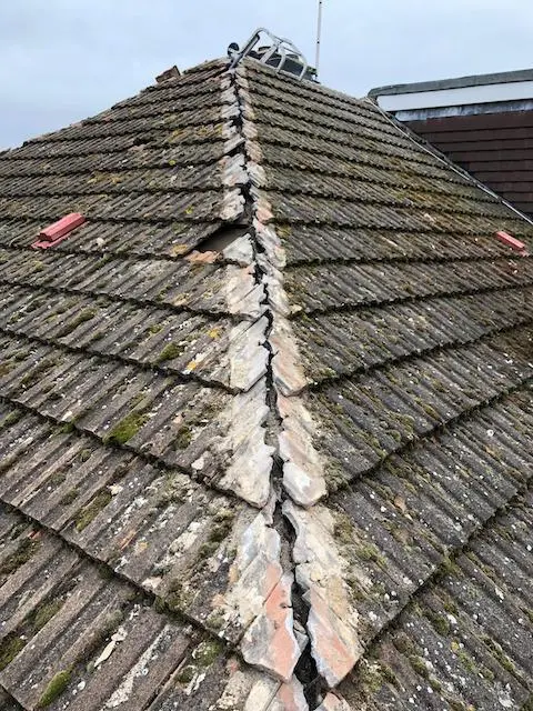Damaged ridge tiles with visible cracks and moss growth on a pitched tiled roof in the UK.