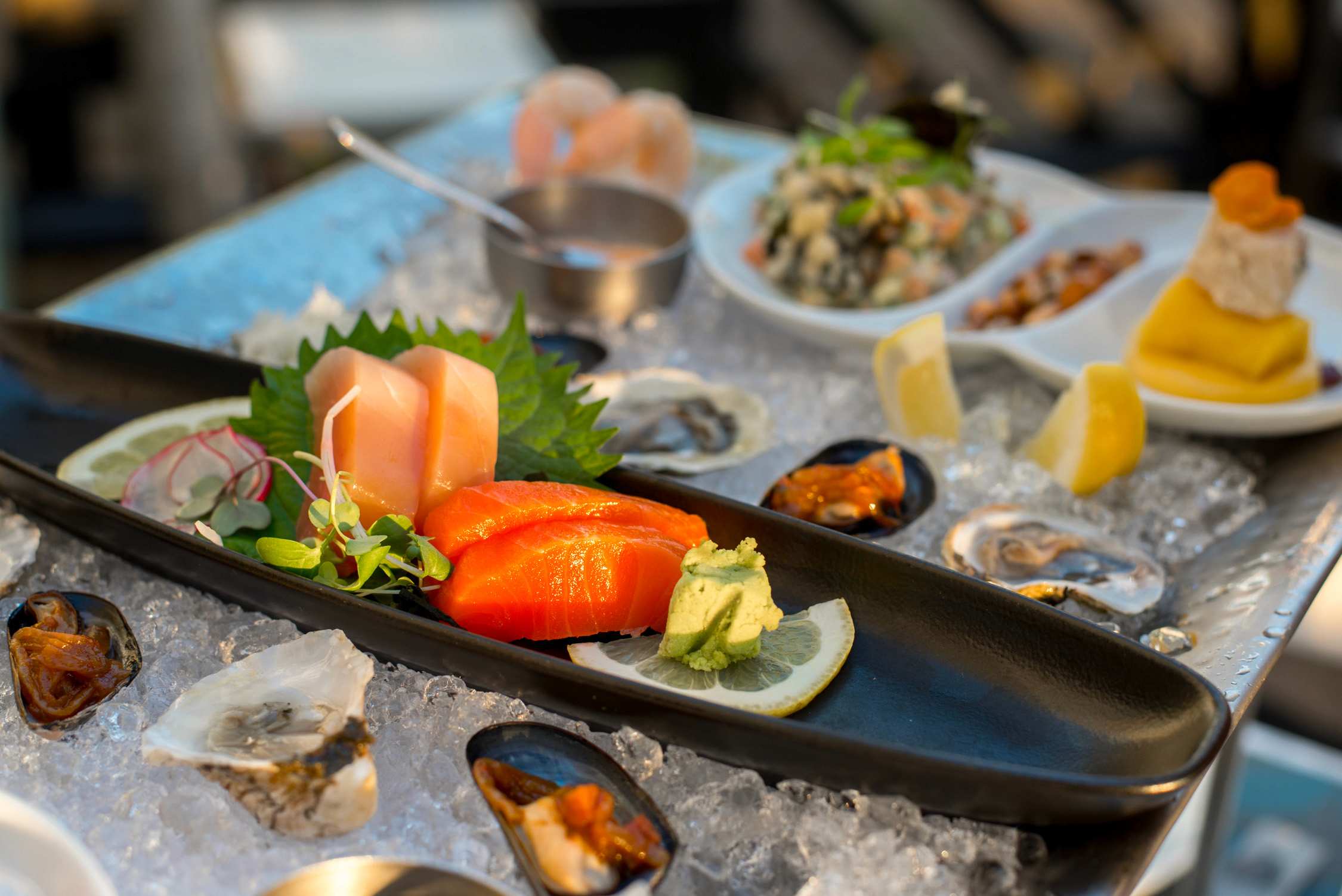 A long black platter and white plates hold assorted sashimi, sushi rolls, and shellfish served on a bed of crushed ice.