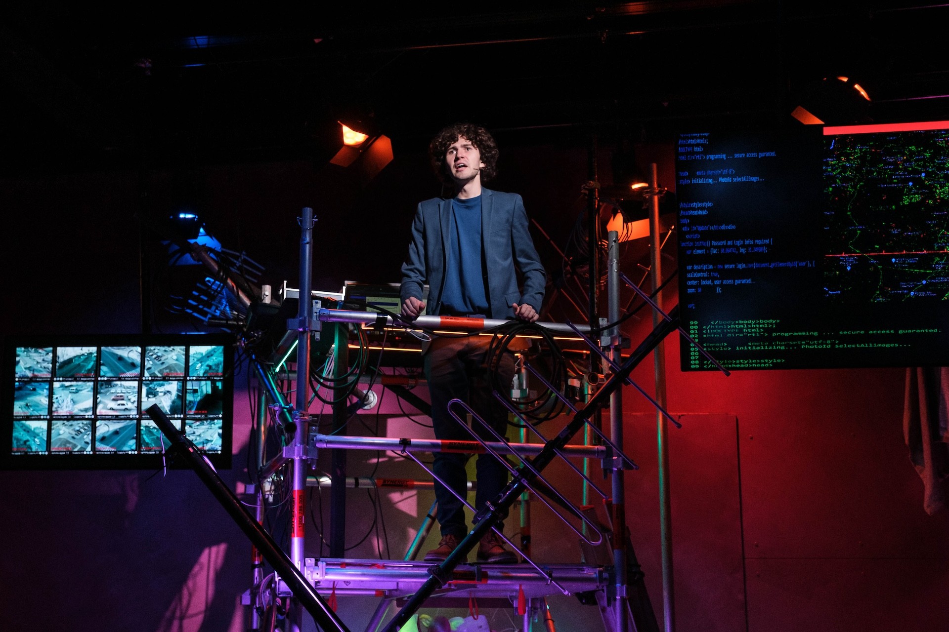 A performer stands atop a scaffold platform surrounded by cables and screens, delivering a speech. One monitor displays surveillance camera grids, while another shows lines of glowing code under vivid stage lighting.