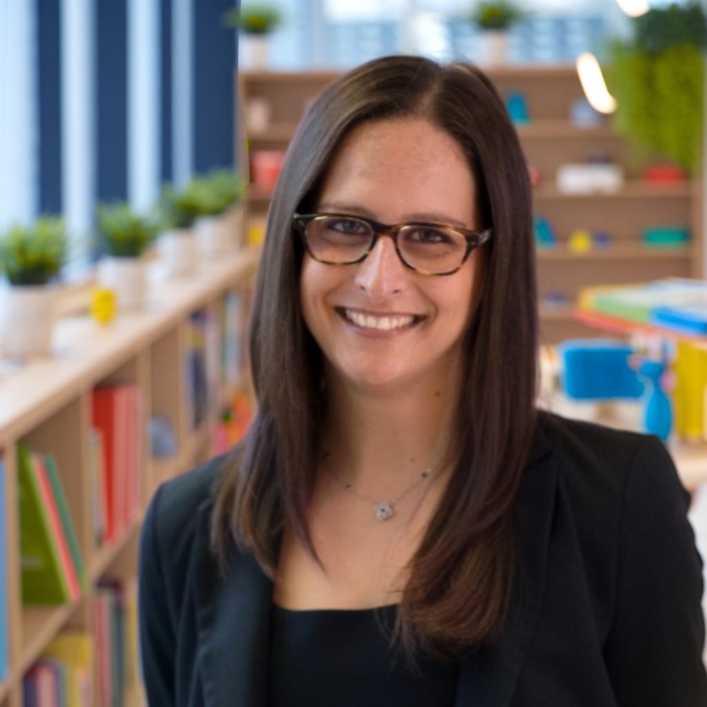 Risa Battino, owner and speech-language pathologist at Communication Connect, smiling in a colorful, book-filled therapy space.