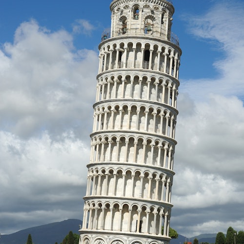 La Torre Pendente di Pisa contro uno sfondo di cielo parzialmente nuvoloso e montagne lontane.