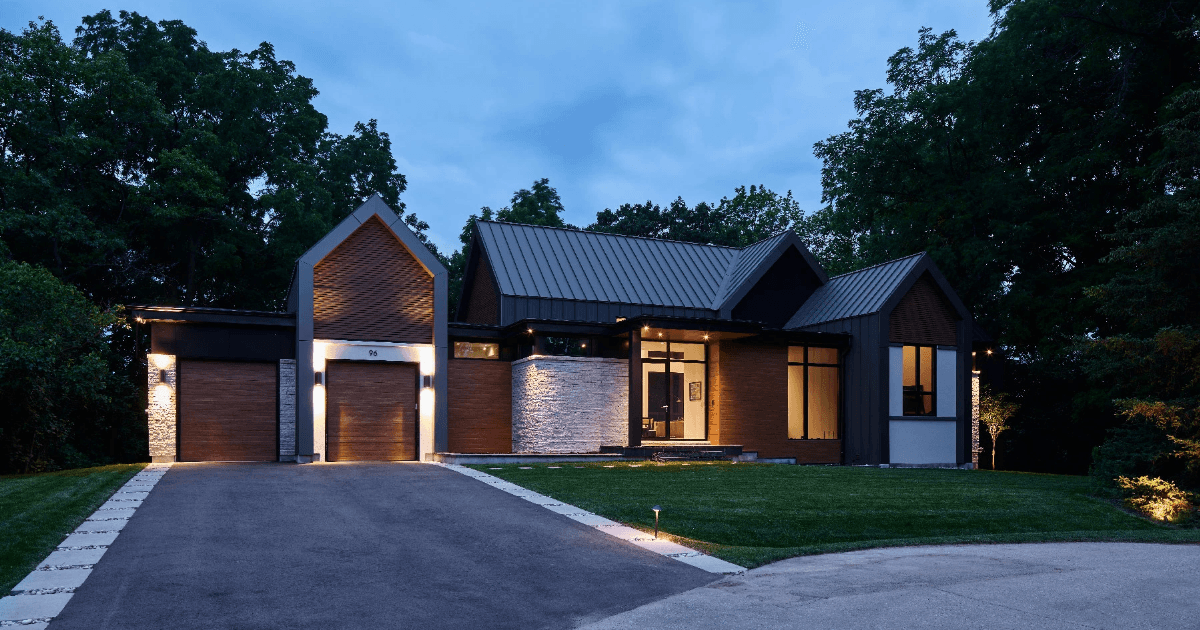 An architectural photograph of a modern, multi-gabled luxury home at twilight. The exterior features a sophisticated mix of dark vertical metal siding, warm wood-toned panels, and textured white stone accents. Integrated LED lighting highlights the clean lines of the roof peaks and the recessed entryway, while large floor-to-ceiling windows reveal a glimpse of a brightly lit, minimalist interior. A smooth asphalt driveway with a decorative stone border leads toward a two-car garage, flanked by a manicured green lawn and dense, dark green trees under a deep blue evening sky.