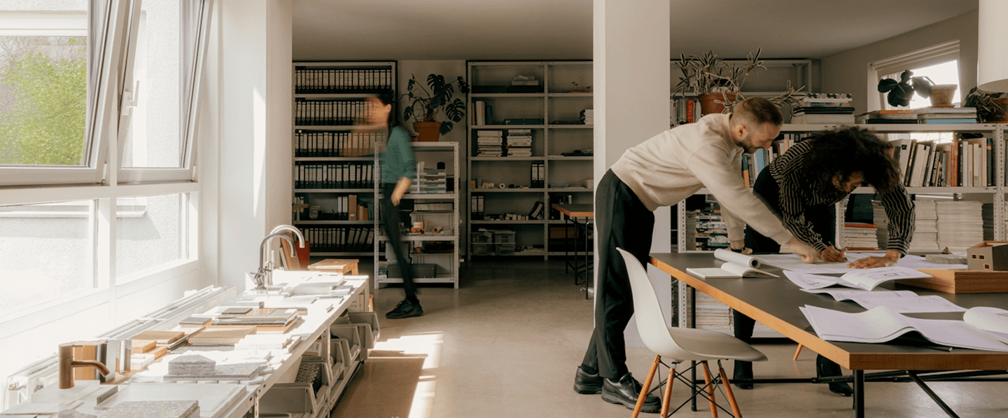 Two architects collaborate on blueprints at a table in a sunny office.