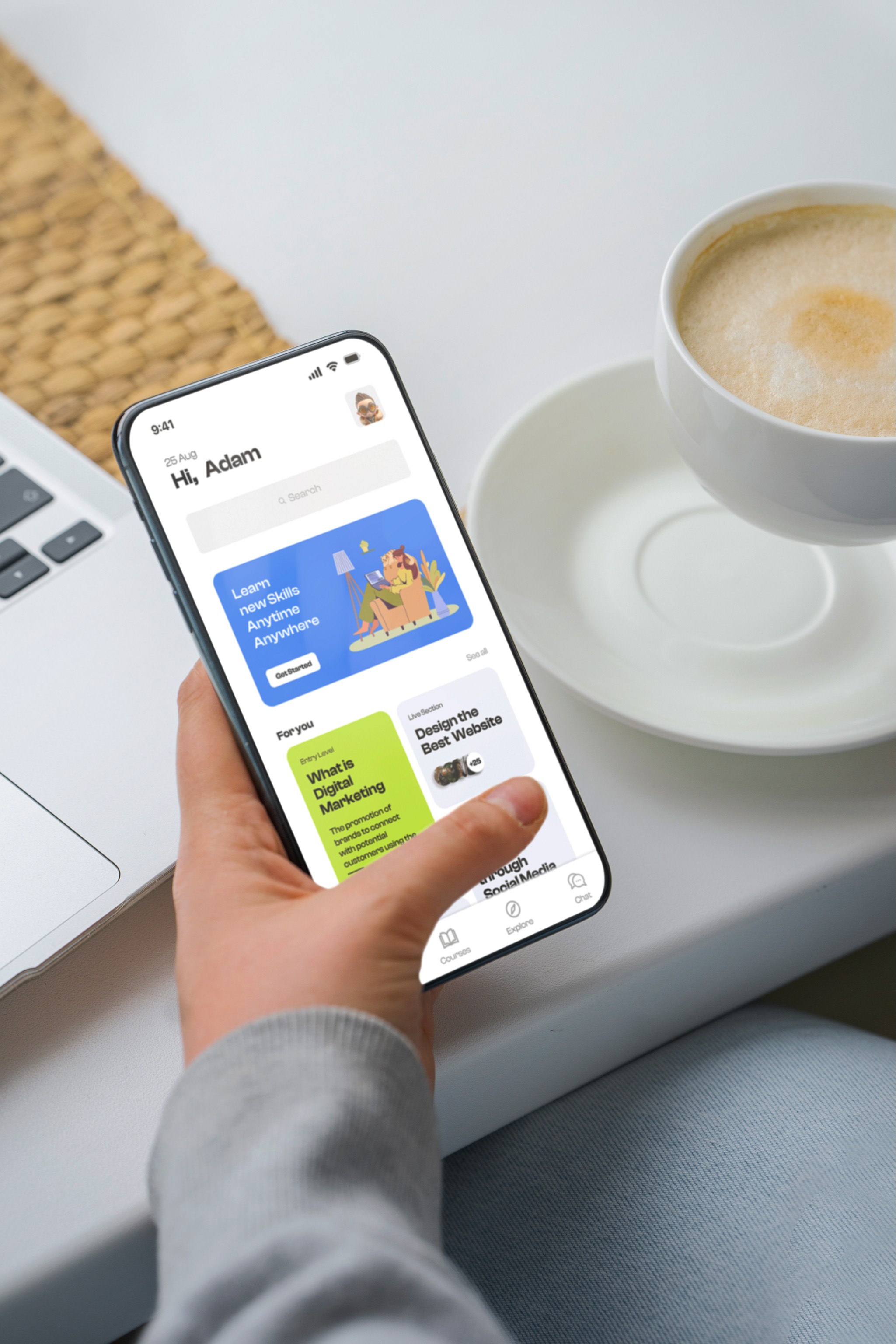 A person is holding a modern smartphone displaying an app with a colorful interface while sitting at a desk next to an open laptop and a cup of cappuccino.