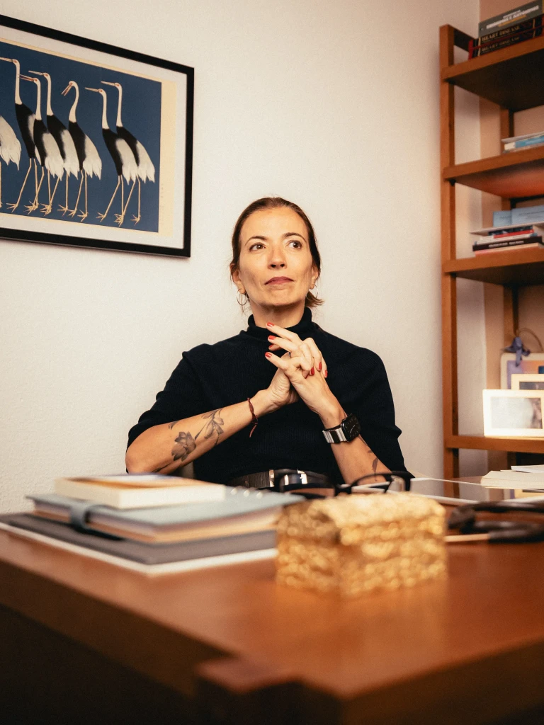 Doutora Ana Cláudia Quintana Arantes, embaixadora da Voa, médica geriatra e paliativista sentada em uma mesa de escritório olhando para o lado e com as mãos entrelaçadas.