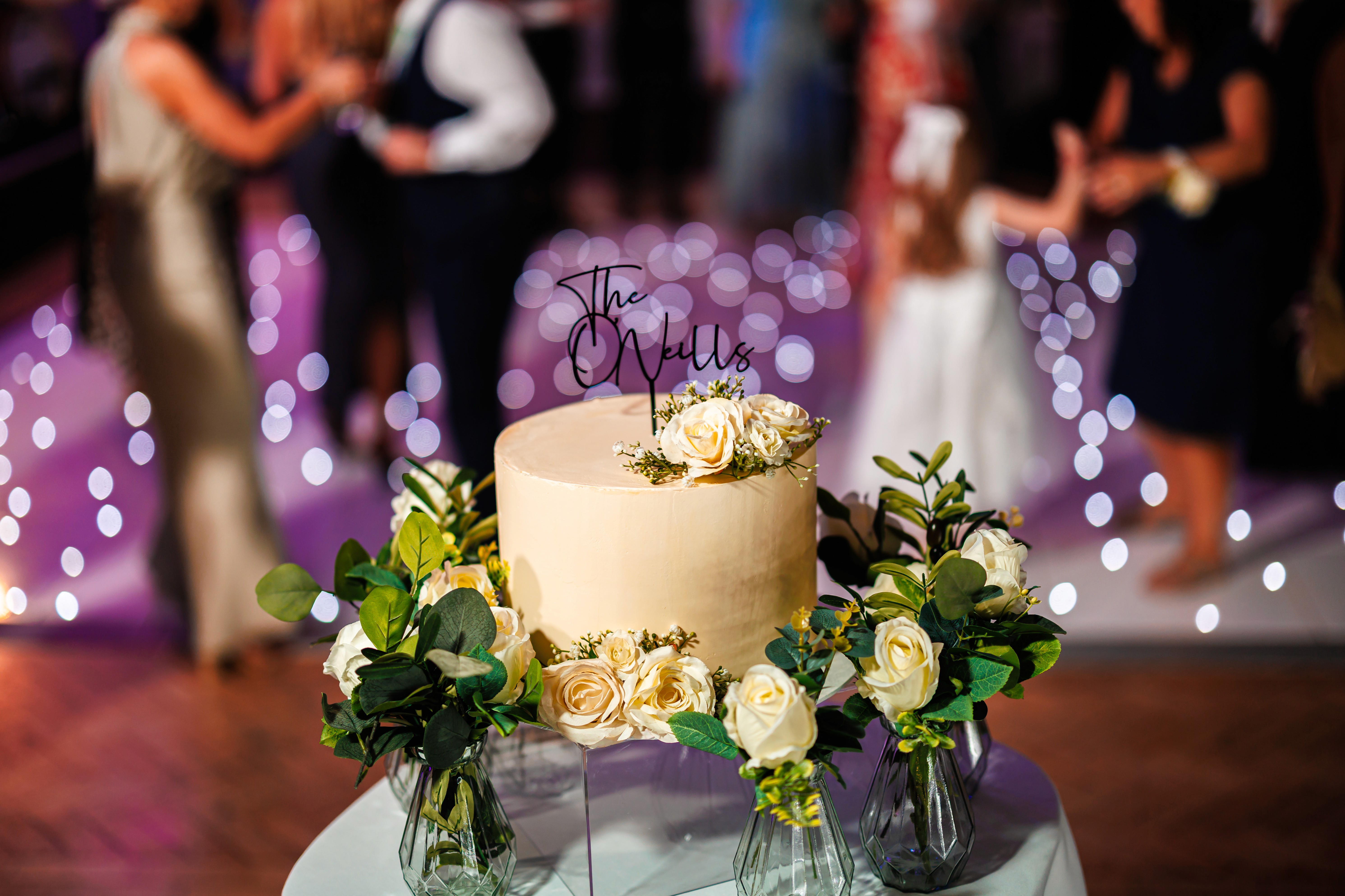 A flower-decorated wedding cake with guests dancing behind it at Mytton Fold