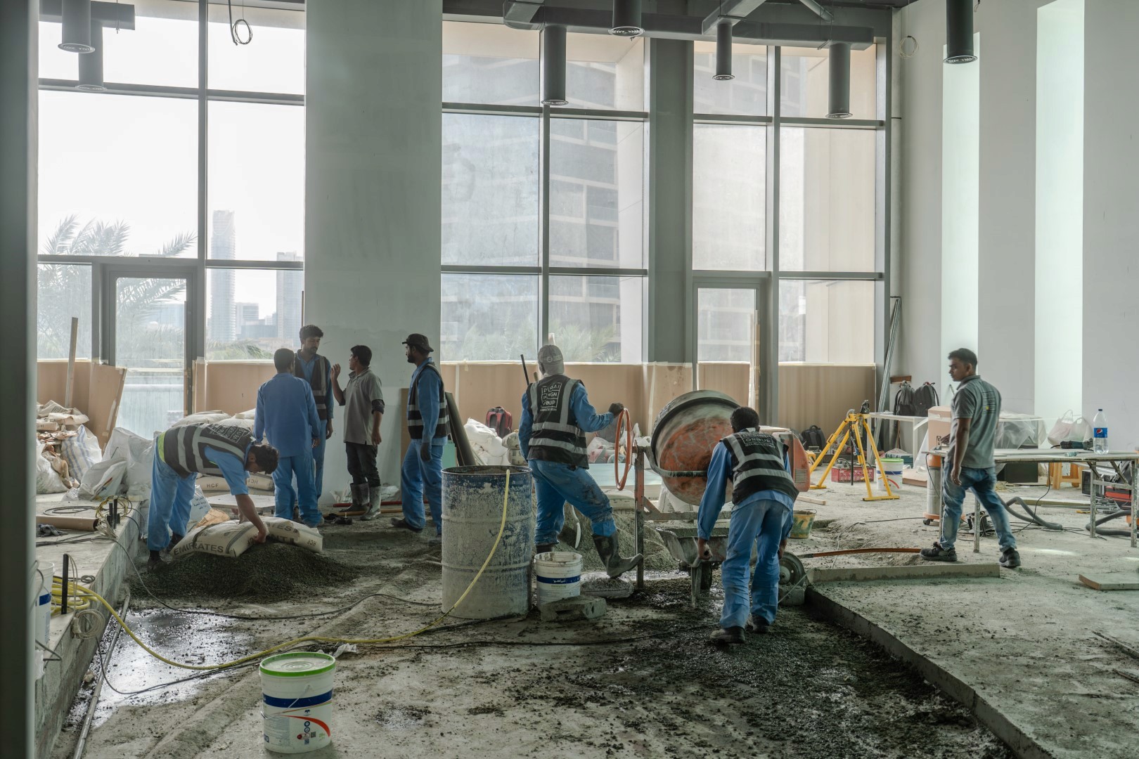A group of construction workers wearing uniforms are actively engaged in pouring and leveling concrete on the floor of a spacious, sunlit building under construction, with large windows and scattered equipment visible in the background.