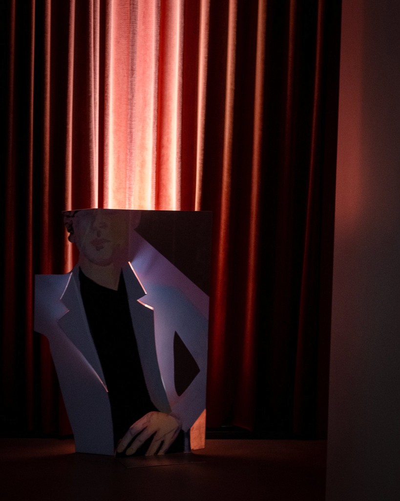 A cardboard cutout of a woman in a white blazer with hands resting on a surface, positioned in front of red curtains with warm lighting.